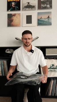 Oggetto nero in una busta di plastica trasparente, tenuto da una persona con una camicia bianca, con dischi e un altoparlante sullo sfondo.