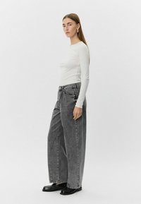 Bela majica z dolgimi rokavi, ohlapni sivi jeans hlače širokega kroja in črne čevlje. Preprost dizajn brez vidnih vzorcev ali okrasitev.