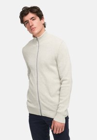 Lichtgrijze zip-up sweater met een textuurpatroon, hoge kraag en geribde mouwen en onderkant. Draagt over donkere broek.
