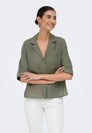 Donna sorridente con capelli scuri raccolti in un basso chignon, indossa una camicia verde a maniche corte con bottoni e pantaloni bianchi, in piedi con le braccia incrociate.