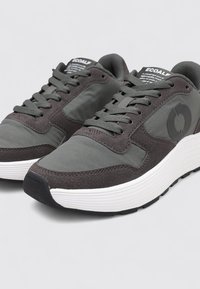 Zapatilla gris con detalles de textil y ante, cuello acolchado, cierre con cordones y una suela gruesa de goma blanca; presenta un detalle de logo en el lateral.