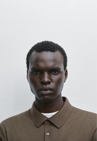 Brown polo shirt with a collar, featuring short sleeves and a subtle texture. Layered over a white shirt, against a plain background.