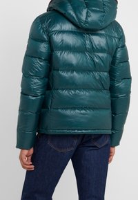 Giacca a vento teal con cappuccio indossata su jeans blu scuro, vista dal retro su uno sfondo chiaro e semplice.