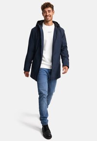 Mørkeblå udendørsjakke med hætte, frontlynlås og sidelommer, lagdelt over en hvid sweatshirt og lyseblå jeans. Sorte sko.