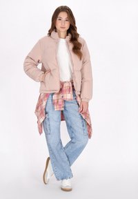 Chaqueta acolchada en rosa claro, con un cuello alto y puños elásticos. Combinada con una camiseta blanca, una camisa de cuadros atada a la cintura y jeans anchos.