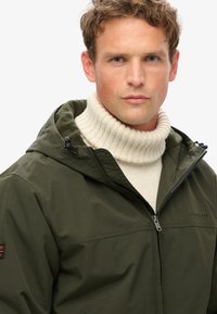 Herre jakke i olivengrøn, vandtæt med lynlåslukning foran, justerbar hætte og tekstureret stof, lagdelt over en cremefarvet ribbet turtleneck sweater.