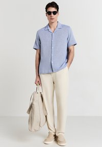 Uomo che indossa occhiali da sole neri, camicia blu a maniche corte con bottoni, pantaloni crema, scarpe beige slip-on, mentre tiene una borsa tote beige.