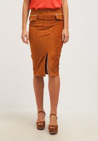 Kvinna som bär en rostfärgad mocka pennkjol med frontslits och fickor med dragkedja, ihop med matchande plattformsklackar.