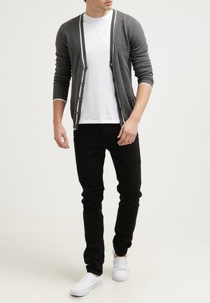 Mies, joka pukeutunut harmaaseen neulotuun napitettuun neuletakkiin valkoisen t-paidan, mustien housujen ja valkoisten slip-on-kengien kanssa, seisoo yksiväristä taustaa vasten.