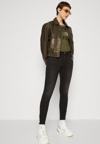 Hnědá kožená bomber bunda s riblovaným lemem, zelené grafické tílko, černé skinny džíny a robustní bílé tenisky.