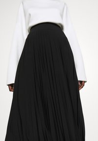 Black pleated skirt with a high waistband, made from a smooth material. A white, long-sleeve top partially visible above the skirt.
