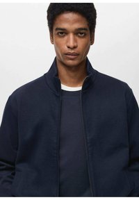 Giacca zip-up blu navy realizzata in tessuto liscio, con colletto alto e tasche laterali, indossata sopra una camicia scura. Design minimalista.