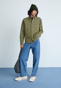 Groene geruite overhemd, lichtblauwe spijkerbroek, witte sneakers met rode accenten en een groene beanie. Houdt een gestreepte tas vast.