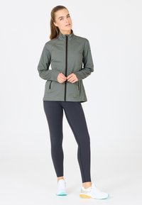 Lichtgewicht, olijfgroene sportjack met een hoge kraag en ritssluiting, gecombineerd met donkere leggings en witte sneakers met kleuraccenten.