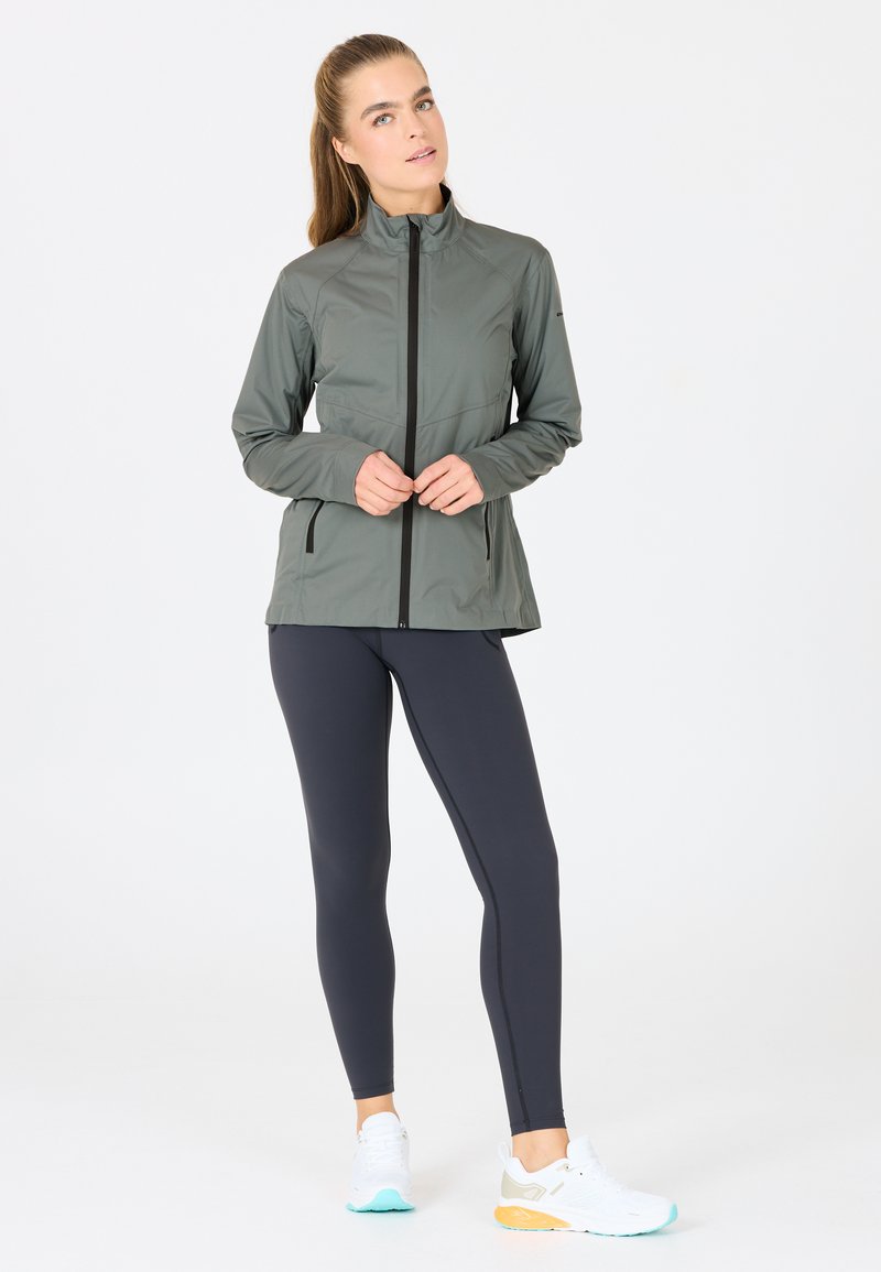 Lichtgewicht, olijfgroene sportjack met een hoge kraag en ritssluiting, gecombineerd met donkere leggings en witte sneakers met kleuraccenten.