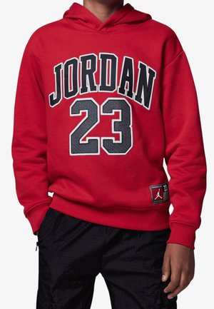 Person iført en rød hættetrøje med "JORDAN 23" i sorte og grå bogstaver og tal, kombineret med sorte bukser.