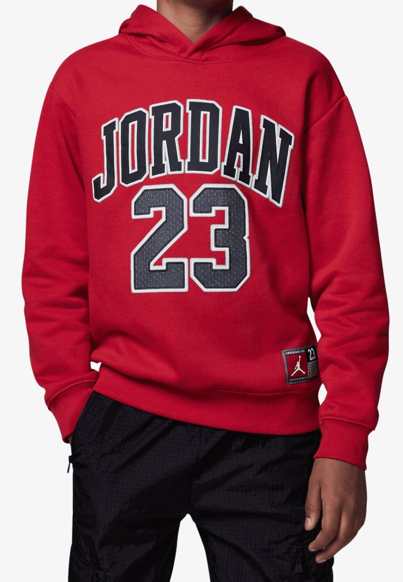 Personne portant un sweat à capuche rouge avec « JORDAN 23 » en lettres et chiffres noirs et gris, associé à un pantalon noir.