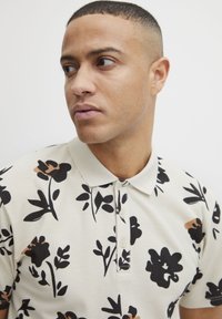 Polo-skjorte i kremfarge med mørkt blomstermønster. Har krage, tre knapper og et strukturert stoff, som gir et avslappet utseende.