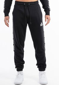Pantalones de chándal negros con cintura ajustable, bolsillos laterales y acentos de logo blanco a lo largo de las costuras exteriores. Tobillos ajustados para un ajuste ceñido.