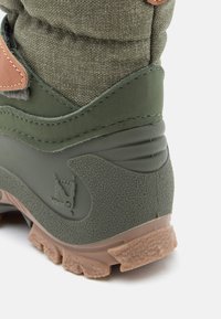 Zöld túracipő, amely texturált felsőrésszel, sima zöld díszítésekkel, barna bőr folttal és strapabíró talppal rendelkezik, amelynek jól meghatározott barázdázása van.