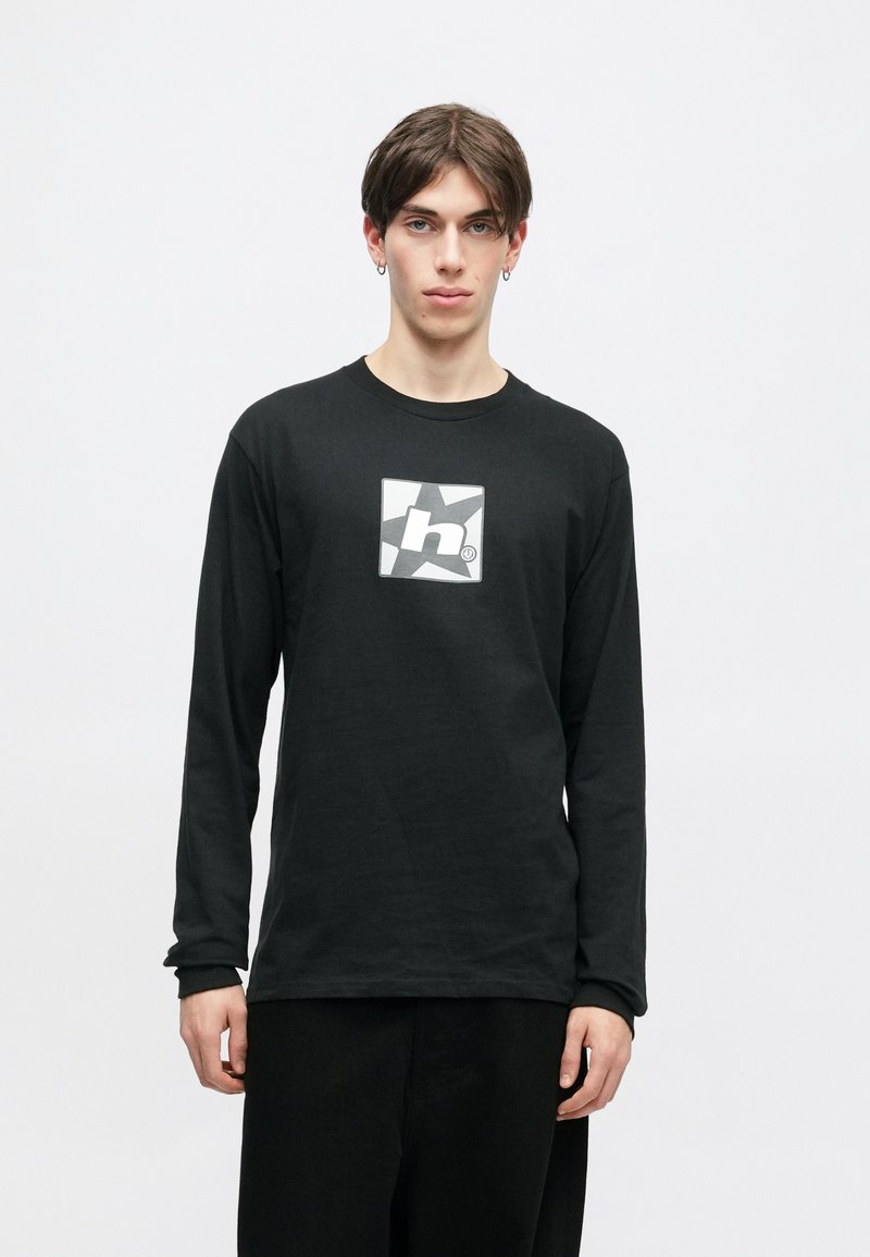 Νέος άνδρας ντυμένος με μαύρο μακρυμάνικο πουκάμισο που έχει λευκό και γκρι λογότυπο αστεριού και ένα μικρό γράμμα "h" στο κέντρο, στέκεται μπροστά σε απλό φόντο.