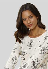 Haut à manches longues, couleur crème, avec des motifs floraux en noir et rose. Il présente un col rond et trois petits boutons à l'avant.