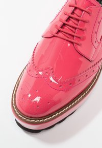 Chaussure brogue rose vif brillante avec perforations décoratives, semelle épaisse marron et rose, et lacets roses assortis.