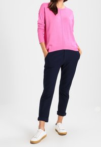 Roze gebreide trui met lange mouwen, gecombineerd met marineblauwe tapered broeken en witte sneakers met bruine zolen. Eenvoudige, casual outfit.