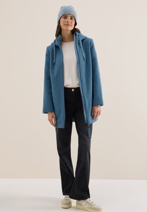Fleece jack in lichtblauw met een volledige rits, trekkoorden aan de capuchon en twee zijzakken. Gepaard met zwarte broeken en crèmekleurige sneakers.
