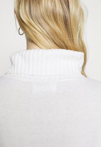 Personne aux cheveux blonds portant un pull à col roulé blanc côtelé, vue de dos avec une boucle d'oreille argentée visible à l'oreille gauche.