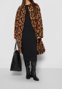 Musterähnlicher brauner und schwarzer Mantel mit markanter "HUGO"-Buchstaben, getragen über einem schwarzen Kleid. Eine schwarze Handtasche und kniehohe schwarze Stiefel runden das Outfit ab.