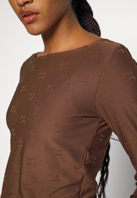 Žena v hnědém dlouhém rukávu s opakujícími se logy Nike Air, s pletenými vlasy staženými dozadu, rty nalíčenými lesklou rtěnkou a malou náušnicí.