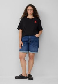 Schwarzes Baumwoll-T-Shirt mit rotem "All Good"-Druck, kombiniert mit blauen Jeansshorts mit umgeschlagenen Säumen. Modell trägt schwarze Schuhe.