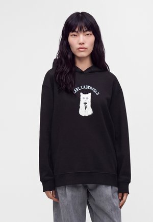 Personne aux longs cheveux noirs porte un sweat à capuche noir avec une illustration de chat blanc et le texte "KARL LAGERFELD", assorti à un pantalon gris.