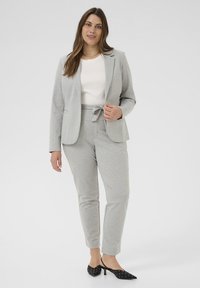 Costume deux pièces gris clair en tissu doux, composé d'un blazer ajusté et de pantalons fuselés avec une taille à nouer. Chaussures à talons noires à pois.