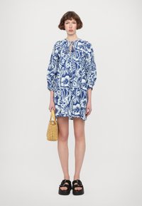 Bloemprint jurk in blauw en wit met een losse pasvorm, pofmouwen en een keyhole halslijn, gecombineerd met een geweven gele tas en chunky zwarte sandalen.