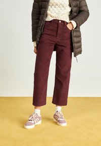 Pantalon en velours côtelé bordeaux à coupe droite, assorti à des baskets rose clair et un pull en maille crème sous une doudoune marron.