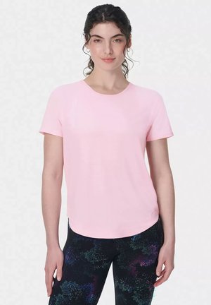 Vrouw met donker krullend haar, gekleed in een lichtroze shirt met korte mouwen en zwarte legging met patroon, staand tegen een effen achtergrond.