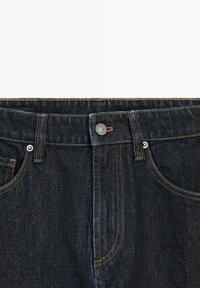 Dunkelblauer Denim-Jeansbund mit Knopfverschluss, Vordertaschen, Gürtelschlaufen und kontrastierender oranger Naht.