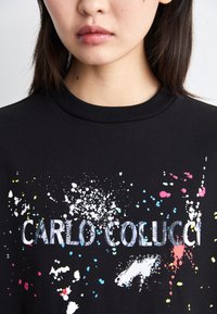 Svart hettegenser med ribbet halslinning, som har flerfargede malingssprut og teksten "CARLO COLUCCI" i et teksturert design.