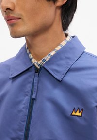 Μπλε φούτερ με φερμουάρ και κοφτή γραβάτα, διαθέτοντας ένα μικρό κεντημένο σχέδιο στέμματος σε πορτοκαλί και μαύρο στο αριστερό στήθος.