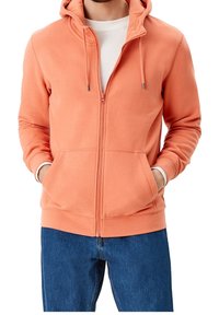 Reißverschluss-Hoodie in Korallenorange aus weichem Stoff. Mit Kapuze, Taschen vorne, Kordelzug und gerippten Bündchen. Kombiniert mit blauen Jeans.