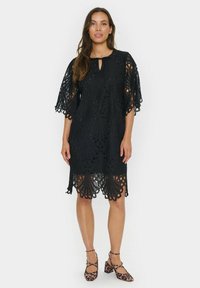 Black lace dress featuring a scalloped hem, floral pattern, and keyhole neckline. Worn with leopard print block-heeled shoes.