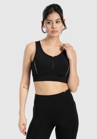 Zwarte sport-bh met een voorrits, brede banden en reflecterende accenten. Gemaakt van een gladde, elastische stof. Gepaard met zwarte high-waisted leggings.