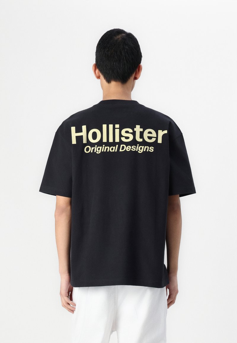 Tricou negru din bumbac, cu un text mare bej pe spate care spune "Hollister Original Designs." Mâneci scurte și croială lejeră.
