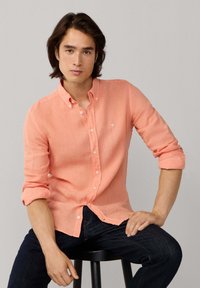 Giovane uomo con capelli scuri di media lunghezza, indossa una camicia salmone abbottonata e jeans scuri, seduto su uno sgabello nero davanti a uno sfondo grigio.