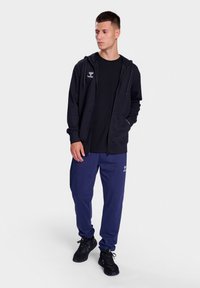 Zwarte zip-up hoodie met trekkoorden, gecombineerd met een zwart t-shirt en marineblauwe joggingsbroek. De schoenen zijn zwart en van sportieve stijl.
