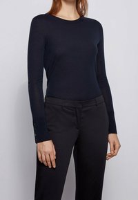 Haut à manches longues bleu marine avec un col rond et une coupe ajustée. Les poignets sont ornés de boutons pour plus de style. Le tissu semble doux et extensible.