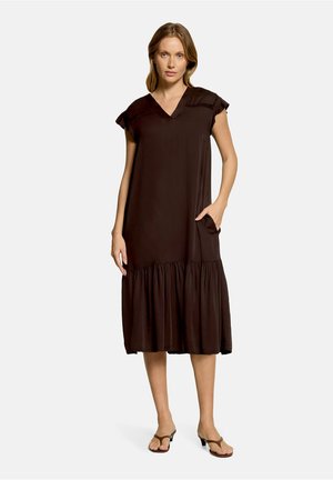 Femme debout portant une robe midi marron foncé sans manches avec un décolleté en V, un ourlet à volants, une poche latérale et des sandales marron à talons.