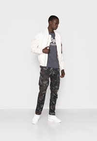 Jachetă bomber albă, tricou grafic gri, pantaloni cargo cu camuflaj și adidași albi; având modele distincte și texturi variate.